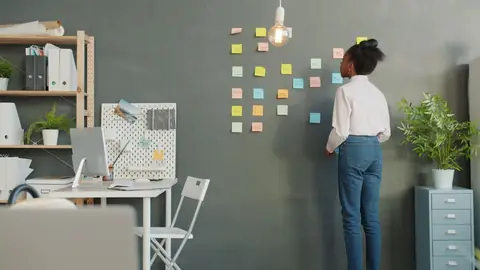 A woman arranges colorful sticky notes on a gray wall in a home office.
