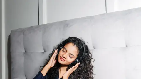 A woman relaxes against a headboard with headphones on and her eyes closed.