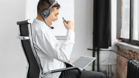 A man wearing a headset talks on a call from an office chair.