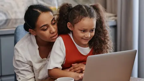 A mother and her young daughter look at a laptop together at home.