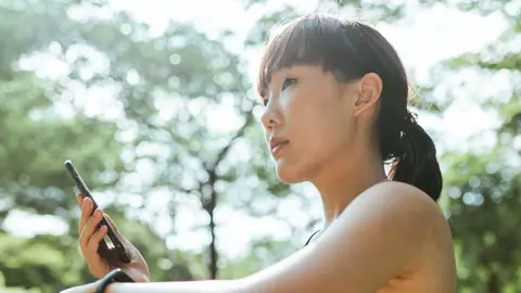 A woman in workout clothes checks her phone outdoors under trees.