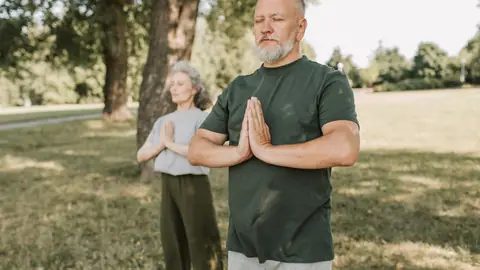 An older man meditates with his palms together in a park, with a woman meditating behind him.