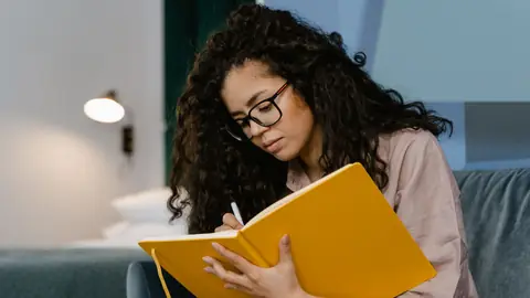 A young woman with glasses writes in a yellow diary on a sofa.