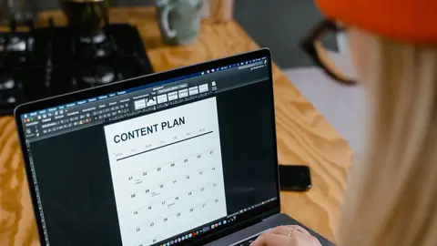 A woman plans a monthly content calendar on her laptop at a wooden desk.