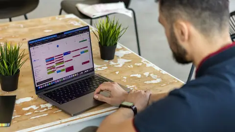 A man works at a cafe table with a color-coded calendar open on his laptop.