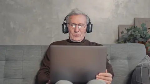 An older man wearing headphones listens while using a laptop on his couch.