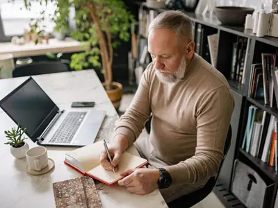 A bearded older man writes in a diary at his home office desk next to an open laptop.