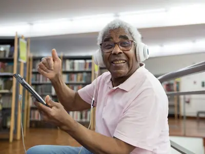 A smiling older man wearing headphones gives a thumbs up to a tablet in a library.