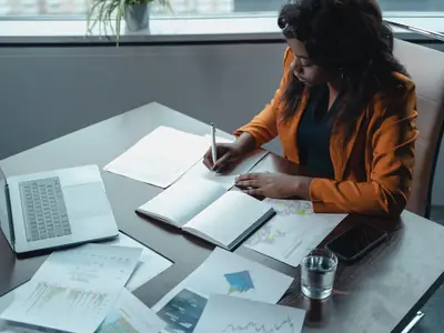 A woman in an orange jacket writes notes at a desk covered in papers, beside a laptop.