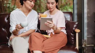 Two East Asian women, one middle-aged and one younger, share coffee and look at a tablet together on a leather sofa.
