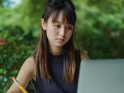 A young woman writes in a notepad while looking at a laptop outdoors.