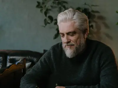 An older man with a gray beard writes on sheets of paper at a dark wooden desk.